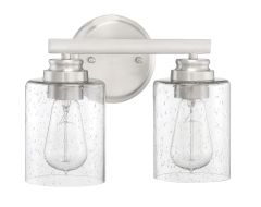 The Bolden - 50502 dual sconce light fixture showcases a brushed metal base with two clear seeded glass shades, each encasing an exposed Edison bulb. Mounted on a circular backplate, it adds elegant charm to any space.